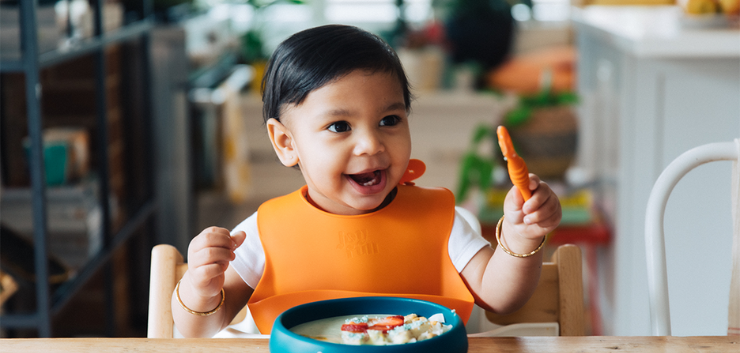 Happy toddler in orange bib seated in high chair about to enjoy his meal.